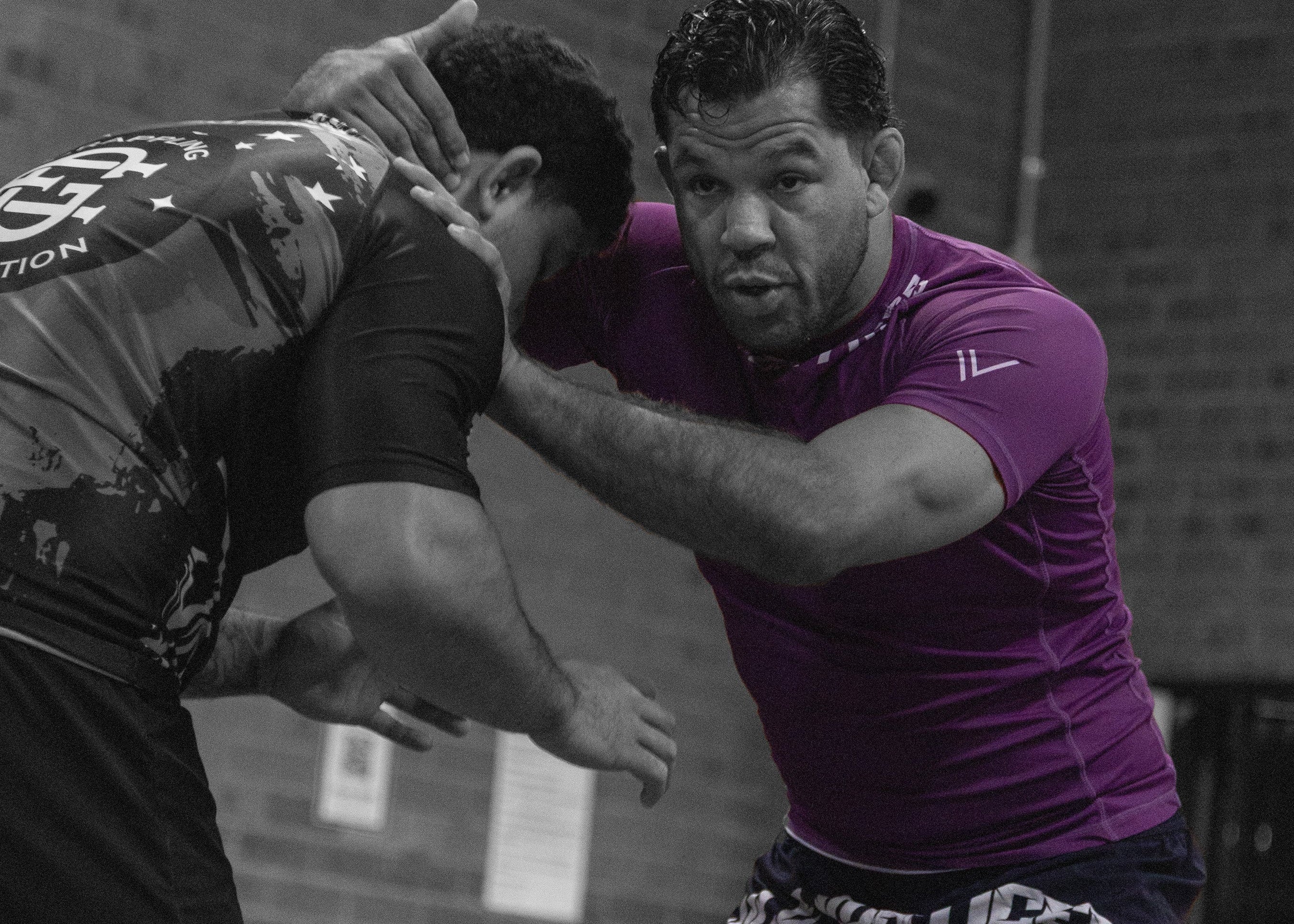 Two men in athletic wear engaged in a physical confrontation, with a brick wall and spectators in the background.
